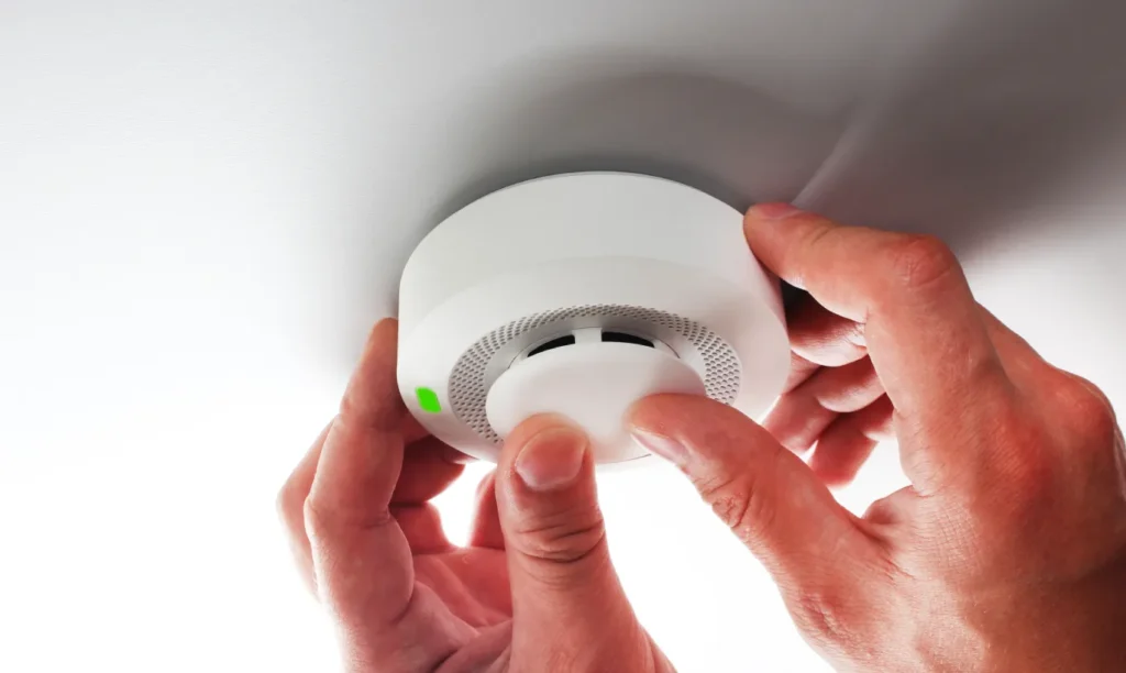 Hands performing fire alarm maintenance by testing a smoke detector mounted on a white ceiling