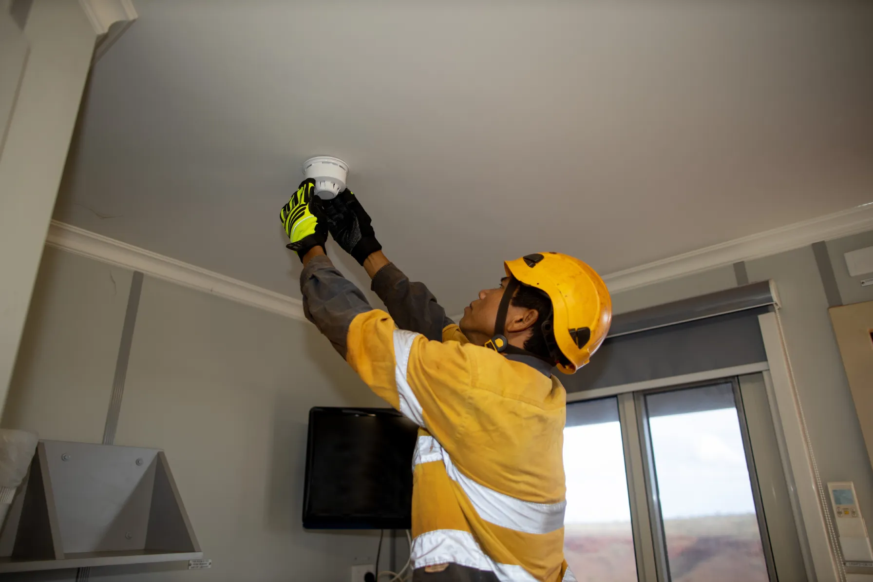 Certified technician in safety gear performing fire alarm maintenance by installing a smoke detector on a ceiling